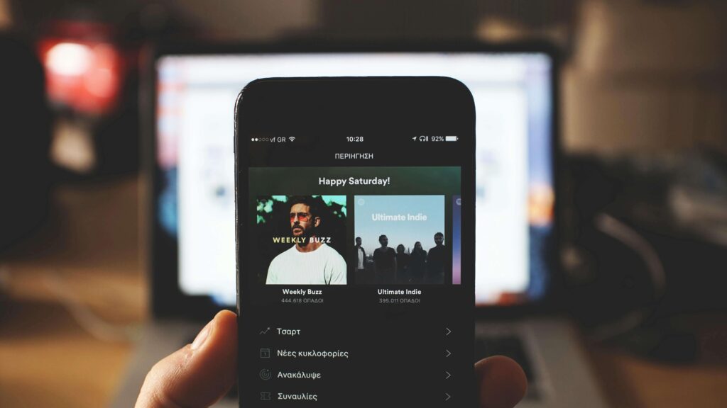 Smartphone exibindo playlists do Spotify, como "Weekly Buzz" e "Ultimate Indie", com um laptop desfocado ao fundo. Técnicas de Neuromarketing do Spotify.